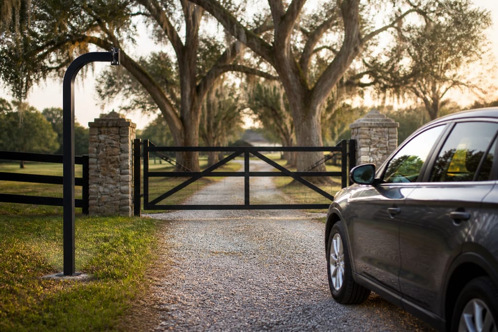 Black GNB-1 gooseneck post on the driver side of a gravel drive, a car length before a ranch gate between stone pillars