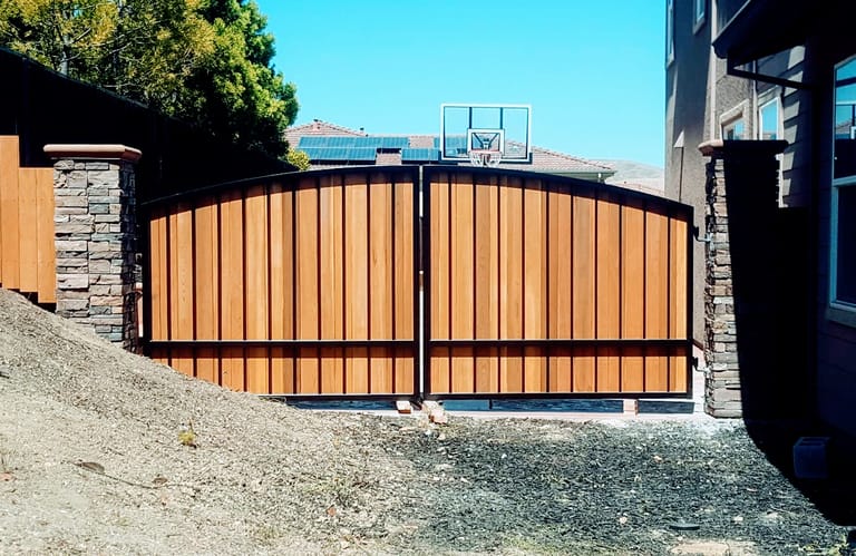 Double swing steel and wood gate hung between stone-clad posts, with ground clearance over a gravel grade