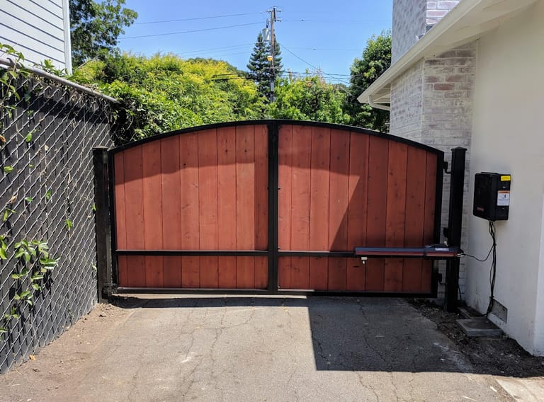 Arched steel-framed double swing gate with wood infill, a swing operator arm and an access-control box on the side wall